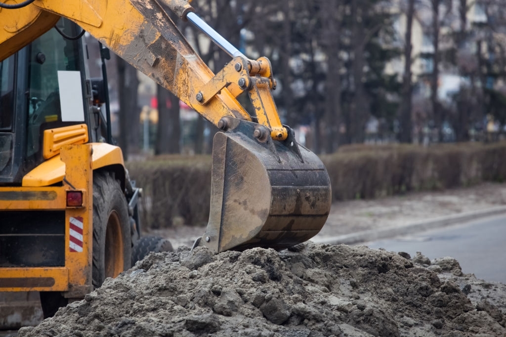 A large yellow excavator stands in the middle of the street near the dug hole.